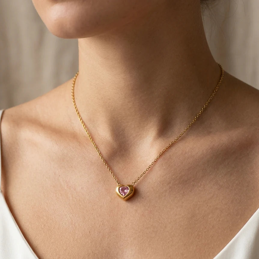 A close-up of a model's neck and chest wearing a delicate gold chain necklace with a small heart-shaped pendant featuring a pink center stone.