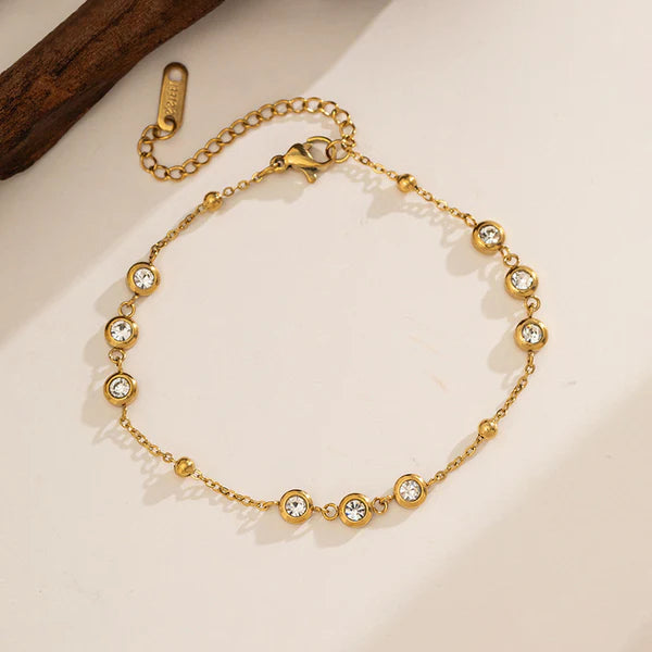 A gold chain bracelet with five bezel-set clear round crystals, displayed on a light beige surface with a dark wood prop in the top-left corner.