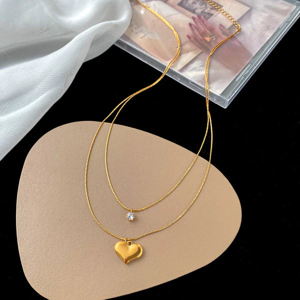 Gold necklace with heart pendant on a beige stand with a magazine in the background