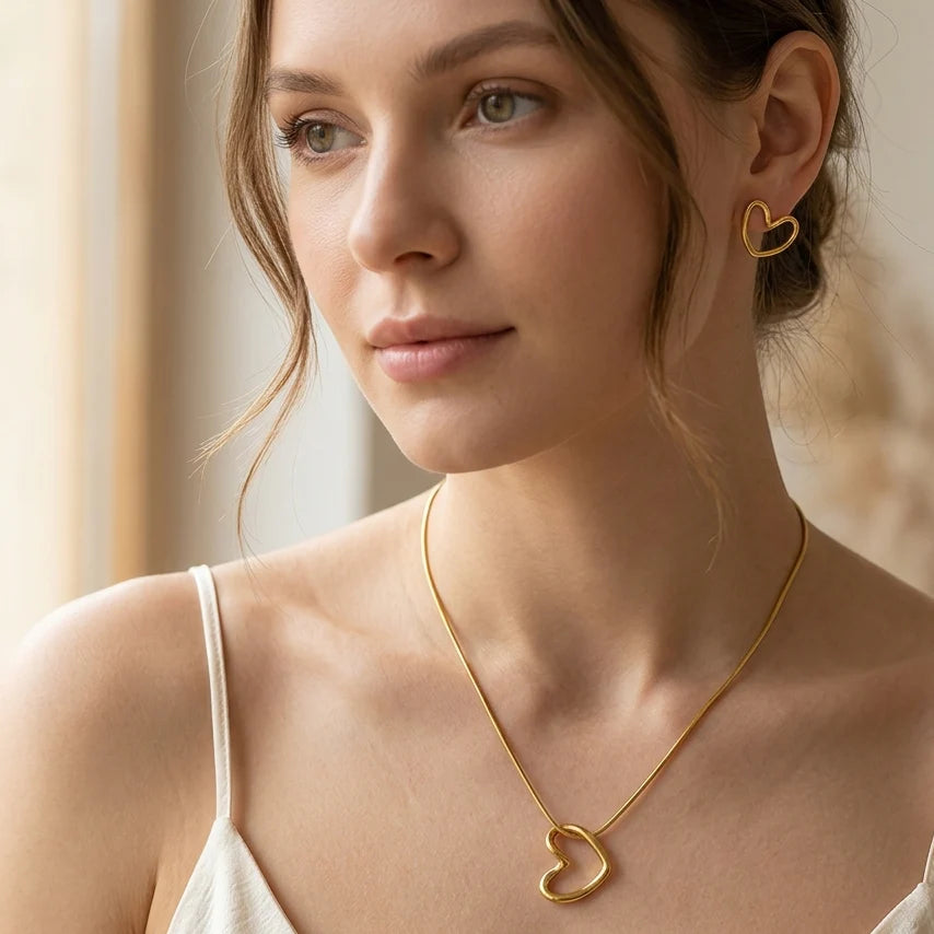 A woman wearing a delicate gold heart-shaped pendant necklace and a matching small gold heart stud earring, shown in a warm, softly lit indoor setting.