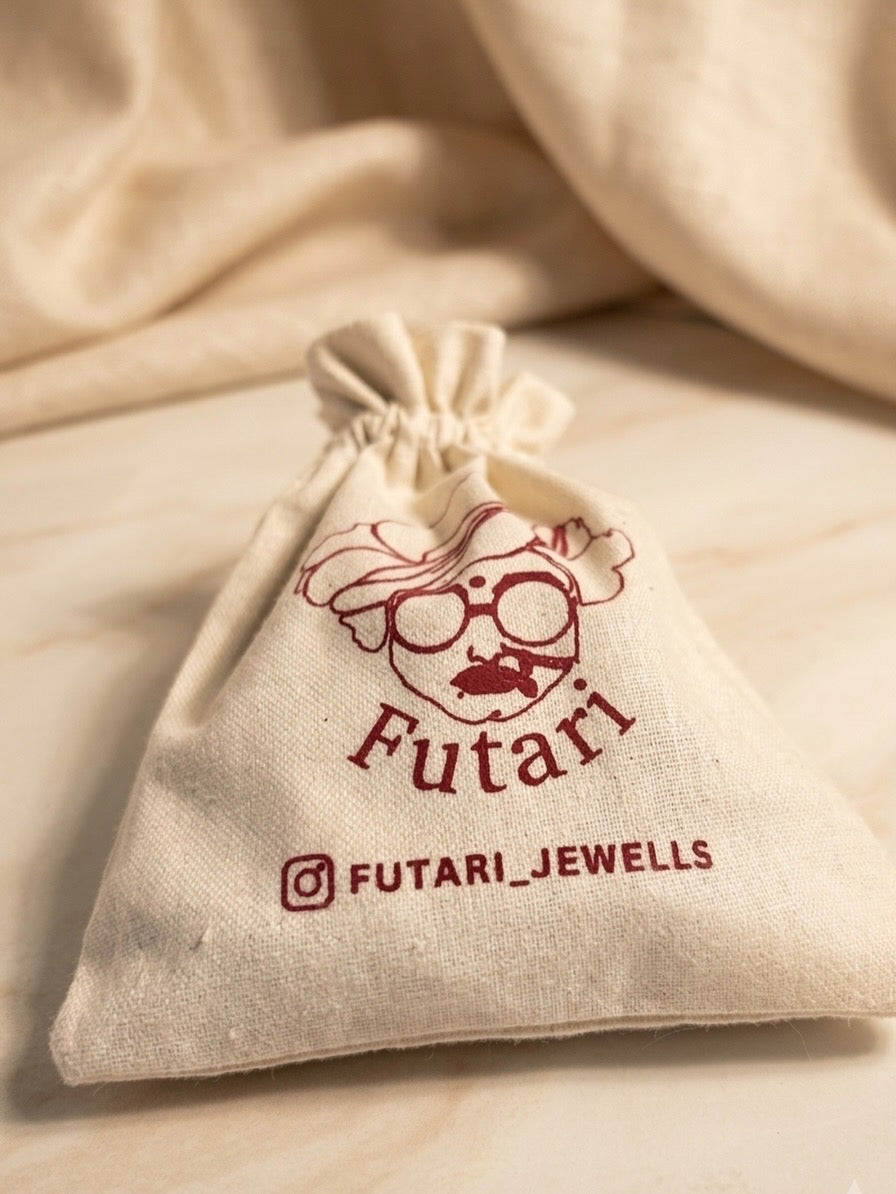 A small beige fabric drawstring pouch with a red printed Futari Jewels logo and handle, resting on a neutral beige surface with draped fabric in the background.