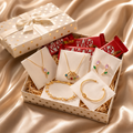 A gift box containing gold jewelry pieces including necklaces, earrings, a bracelet, and a cuff bracelet, arranged on white cards in a straw-lined box. A KitKat bar is visible behind the jewelry, and a matching gift box with gold polka dots sits behind the open box on a champagne satin fabric background.