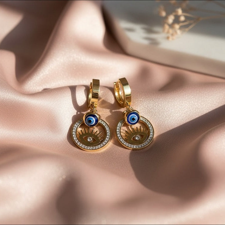 Gold earrings with blue eye design on a pink fabric background