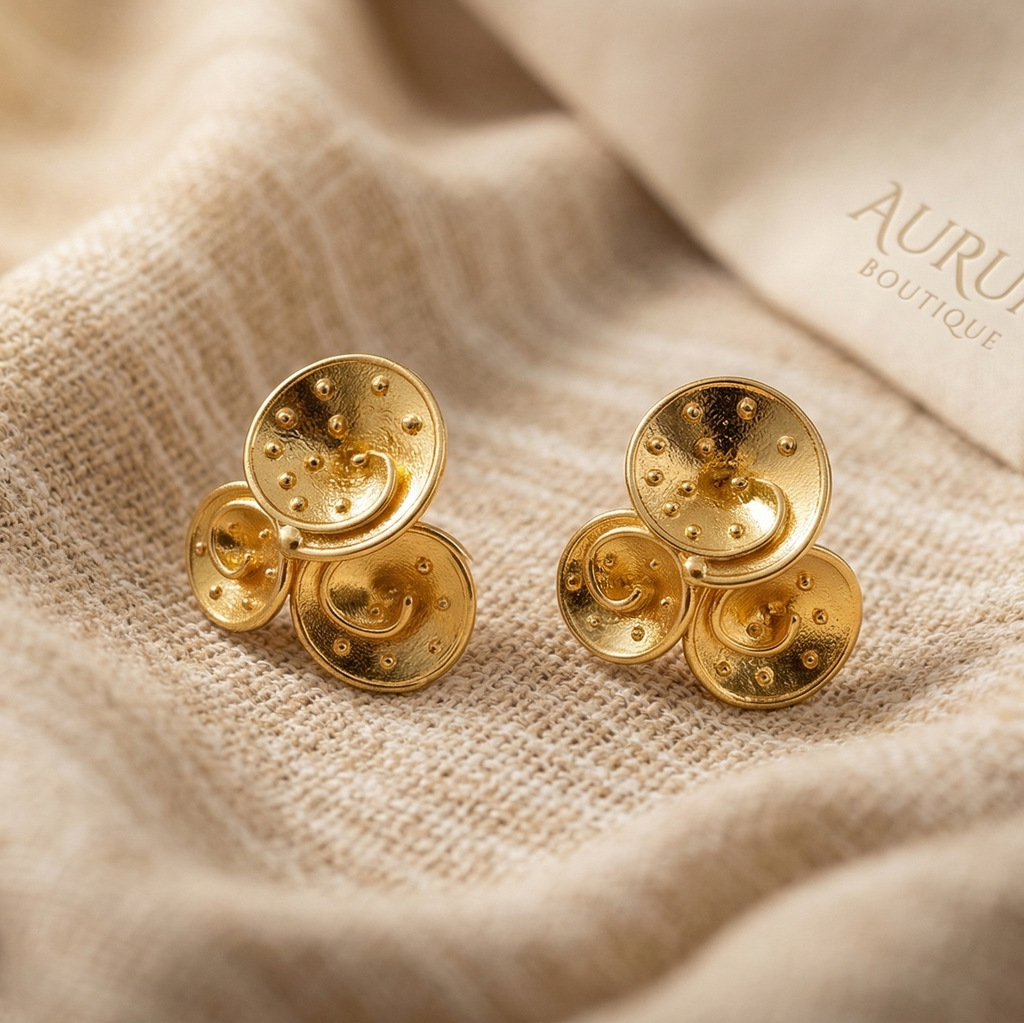 Gold earrings on a textured beige fabric with 'Aurum' branding.