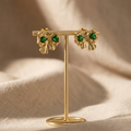 Gold earrings with green gemstones on a gold stand against a beige fabric background