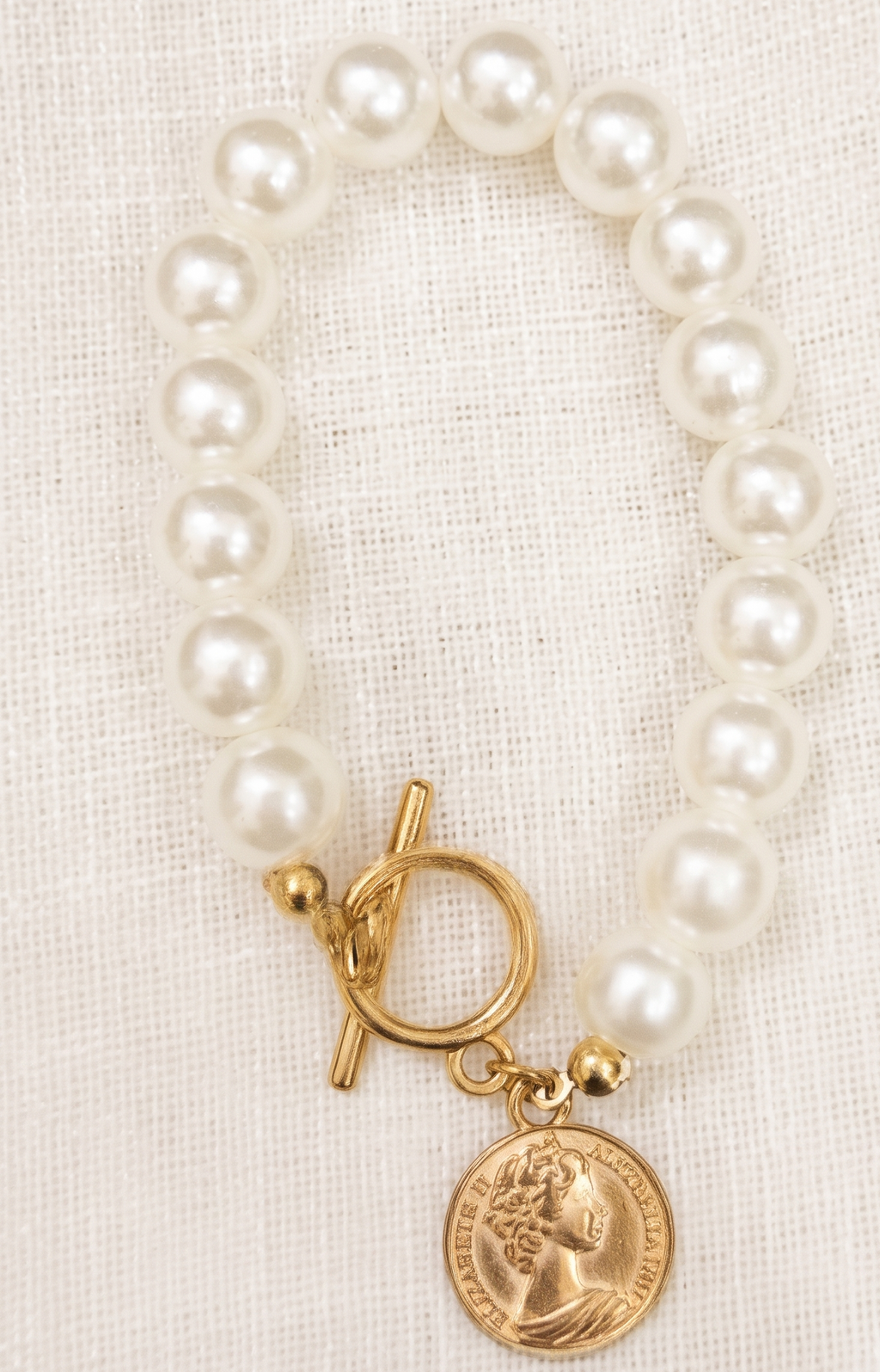 Pearl bracelet with a gold clasp and coin charm on a beige fabric background