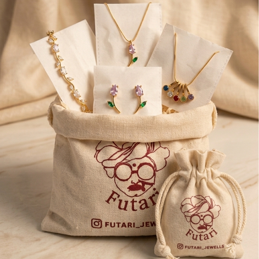 A jewelry gift set displayed in a beige canvas bag with the brand name Futari Jewels printed on it, alongside a matching drawstring pouch. The jewelry set includes gold necklaces, floral stud earrings, and a colorful shell-shaped pendant, all arranged on white display cards against a draped beige fabric background.
