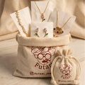 A jewelry gift set displayed in a beige canvas bag with the brand name Futari Jewels printed on it, alongside a matching drawstring pouch. The jewelry set includes gold necklaces, floral stud earrings, and a colorful shell-shaped pendant, all arranged on white display cards against a draped beige fabric background.