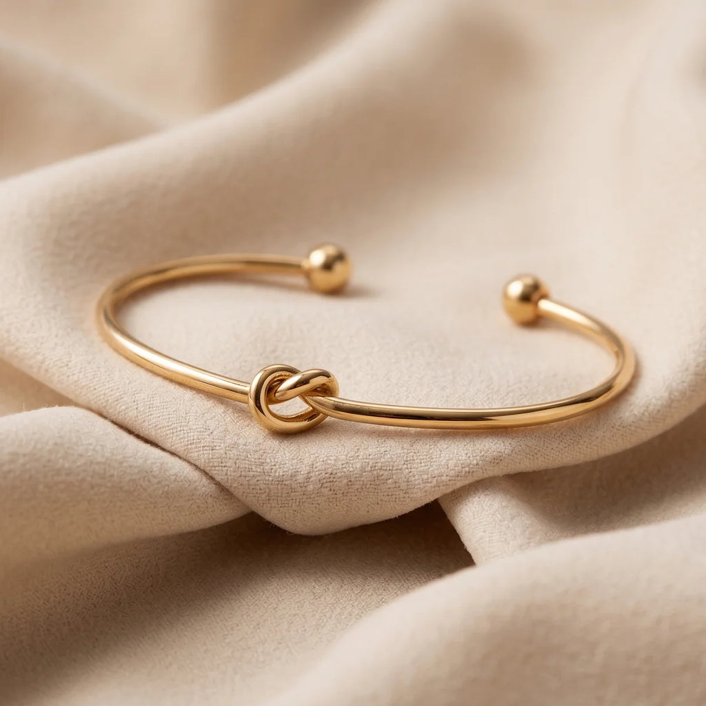 A thin gold open cuff bracelet with a knotted center detail and small bead ends, displayed on draped beige fabric.