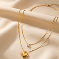 A double-layered gold necklace with a small clear stone pendant on one layer and a polished gold heart pendant on the other, displayed on draped cream-colored satin fabric.