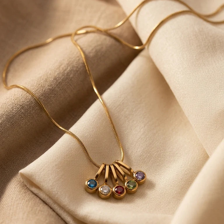 A gold necklace with a multi-stone pendant resting on draped beige fabric. The pendant features a fan-like gold design with five small round bezel-set gemstones in blue, clear, red, green, and purple.