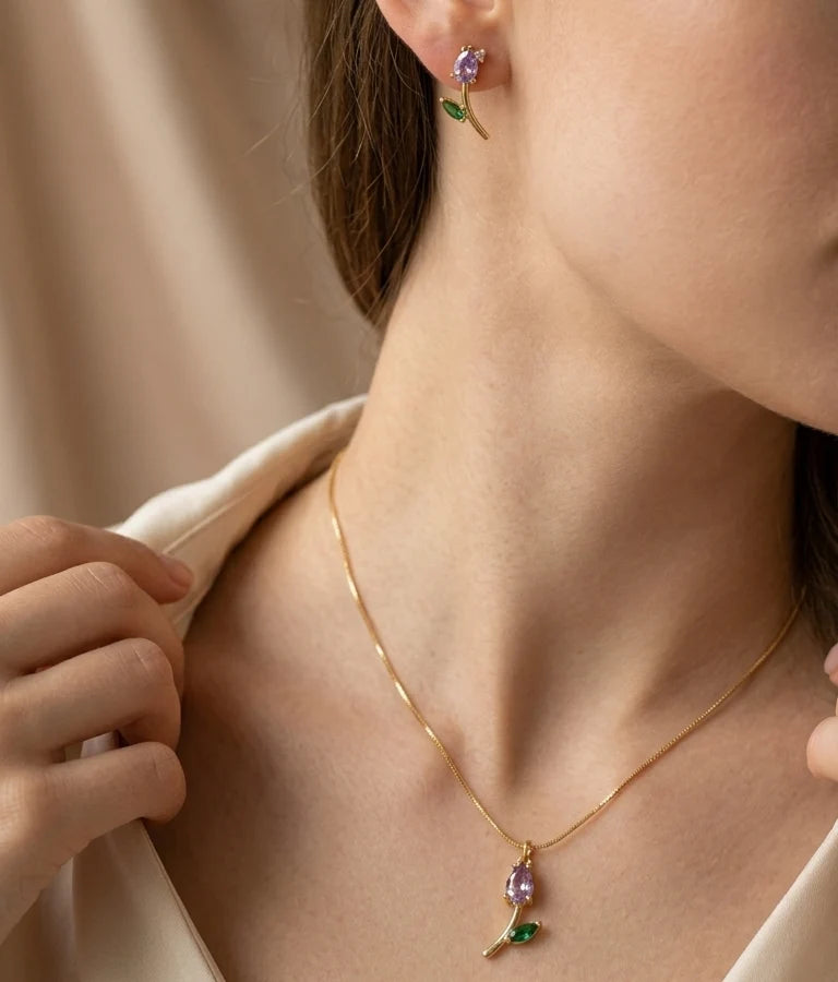 A close-up lifestyle shot of a model wearing a delicate gold necklace and matching stud earring, both featuring a small purple flower and green leaf charm.
