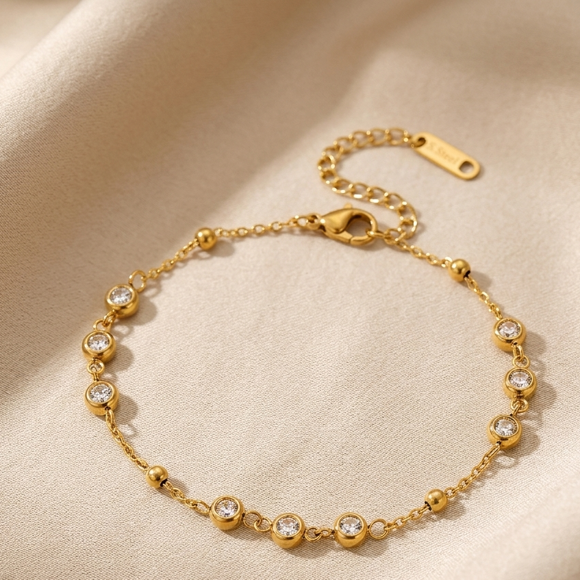 A delicate gold chain bracelet with small bead accents and clear round gemstones, displayed on a soft beige fabric background.
