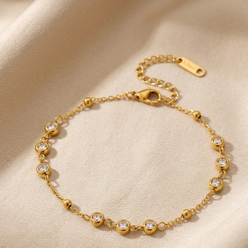 A delicate gold chain bracelet with small bead accents and clear round gemstones, displayed on a soft beige fabric background.