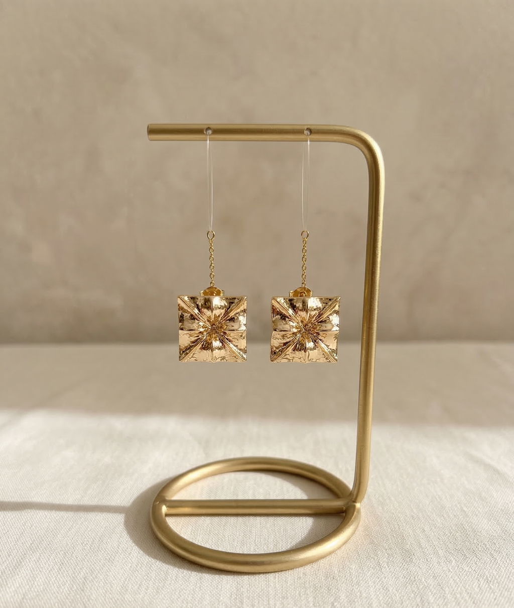 Gold square earrings on a gold stand against a beige background