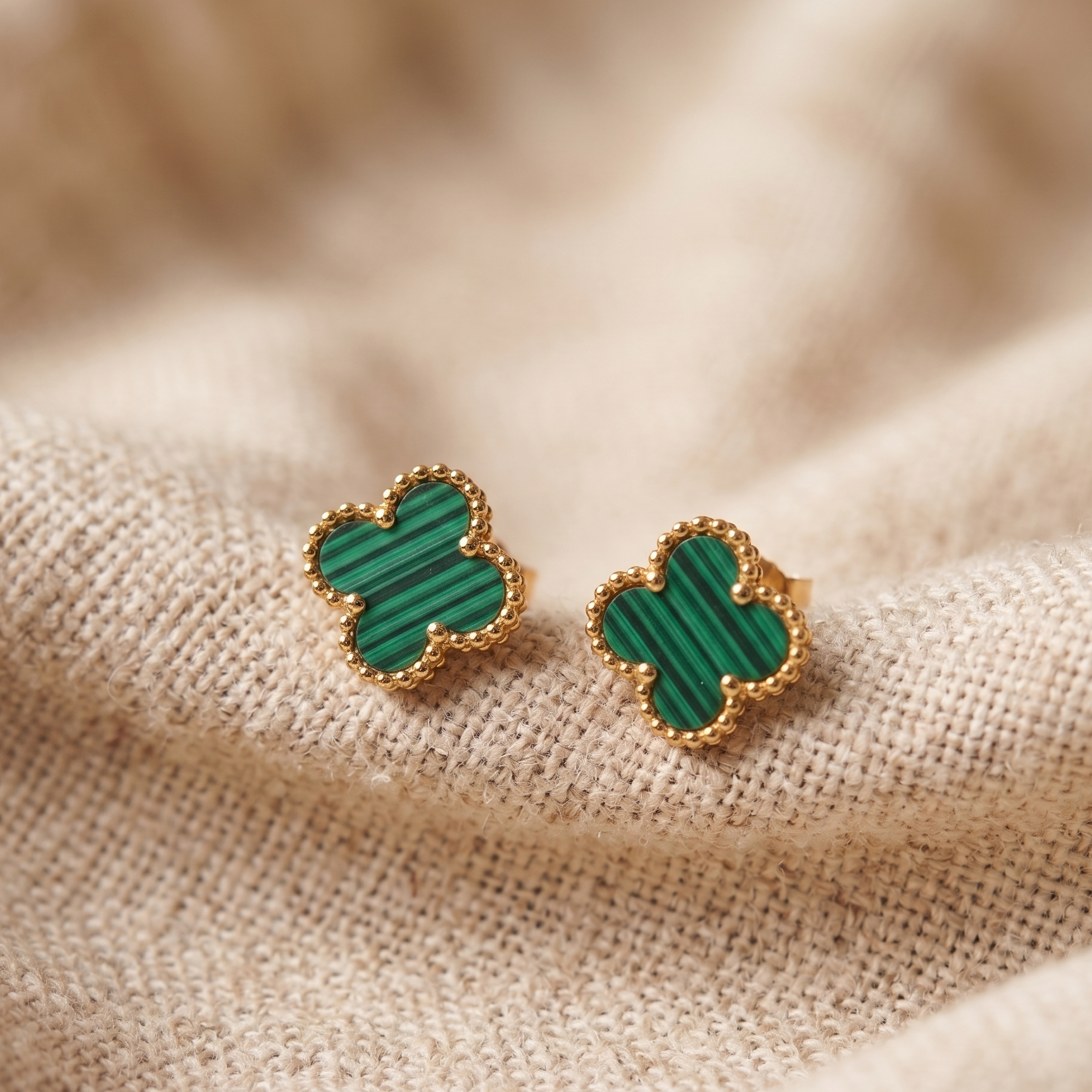 Green clover-shaped earrings with gold accents on a beige fabric background