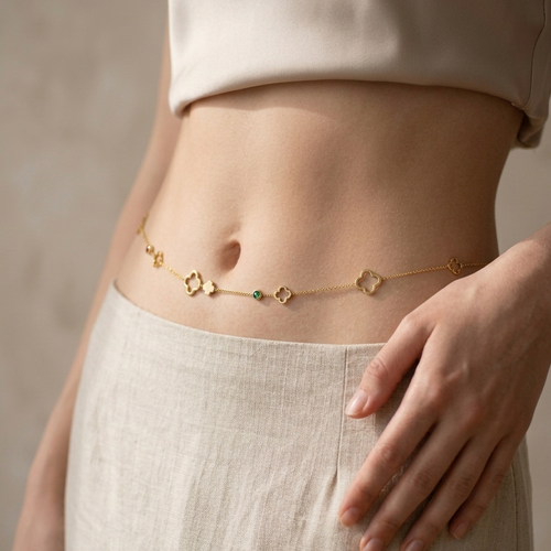 A close-up of a model's waist wearing a delicate gold chain belly chain with small floral and geometric charms, styled with a cream-colored linen top and skirt.