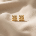 Gold square stud earrings on a textured beige surface