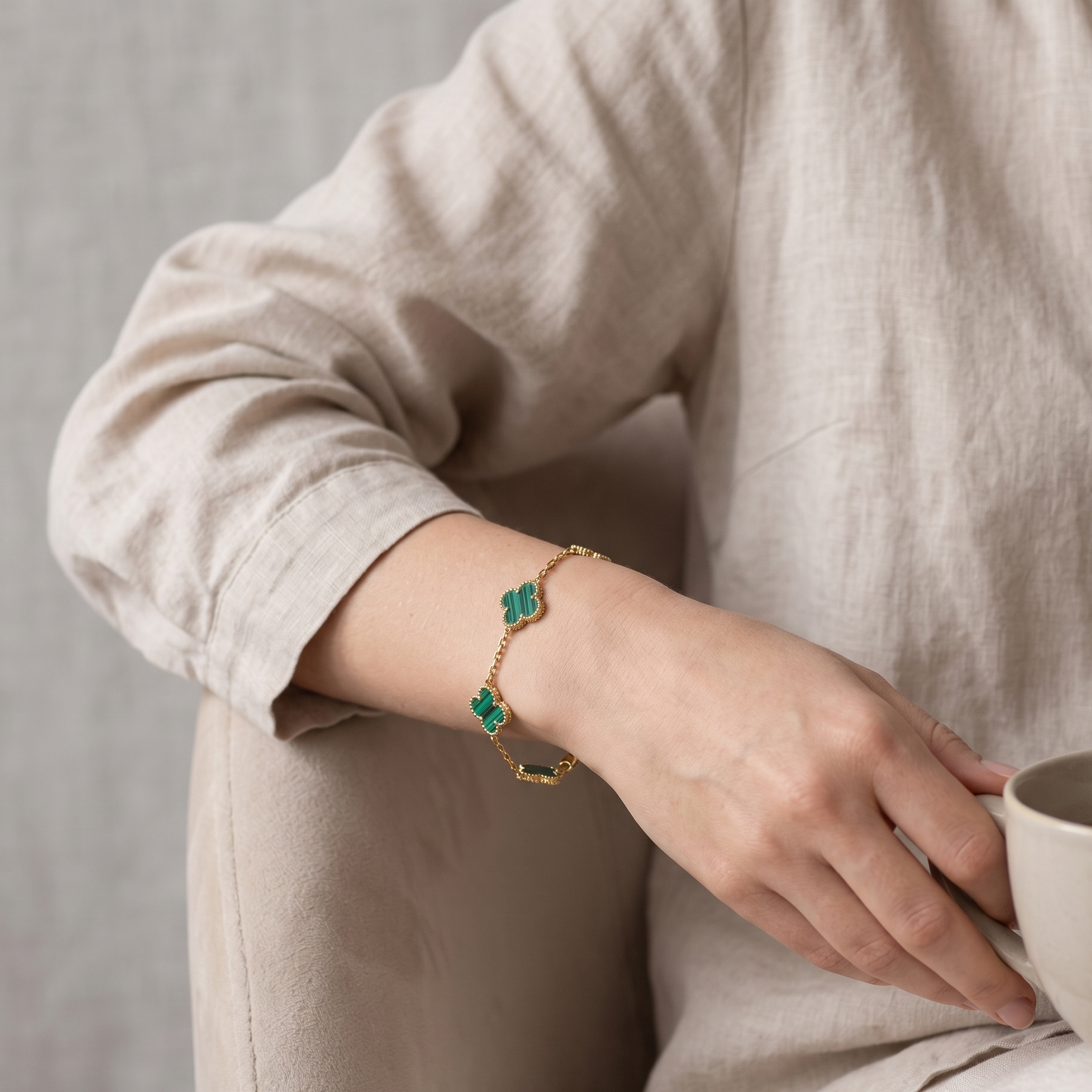 Person wearing a gold bracelet with green stones, holding a cup against a neutral background