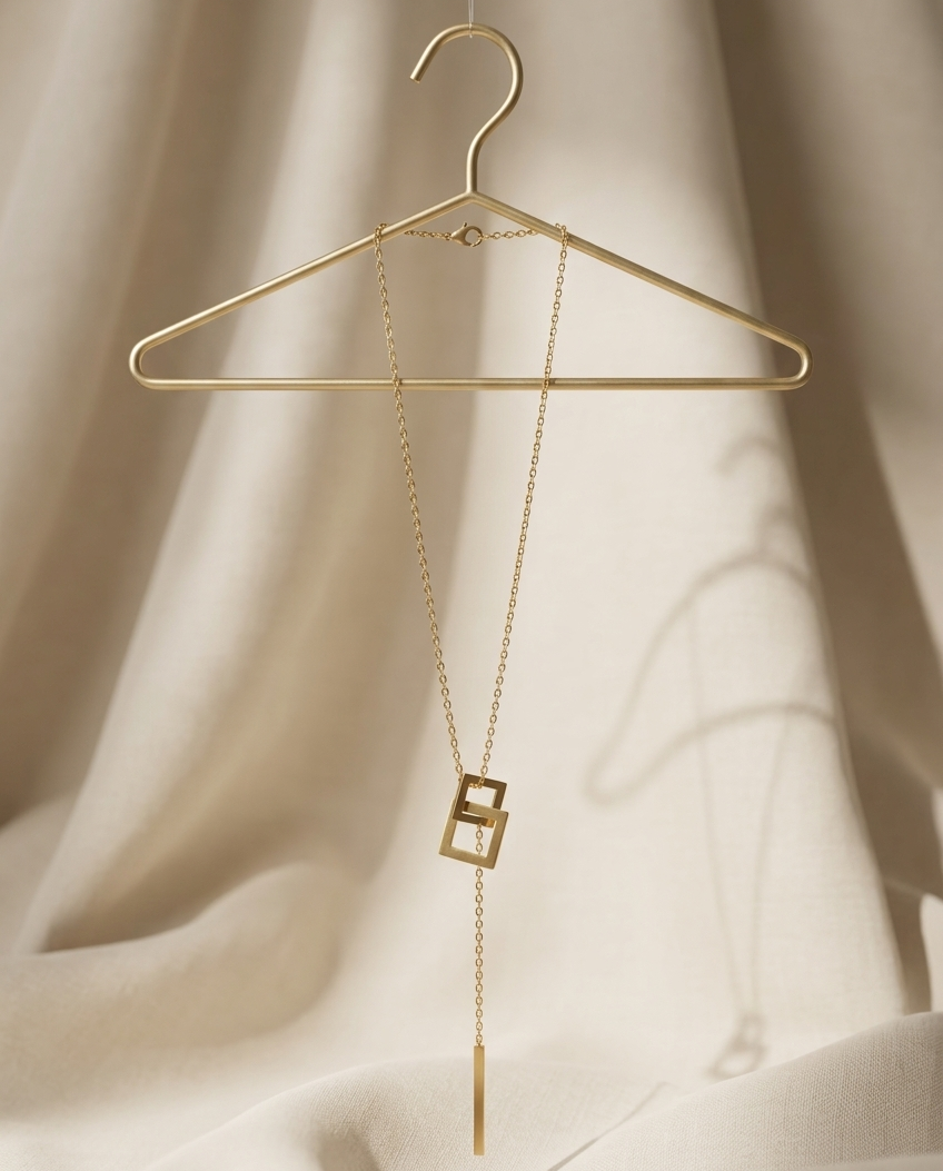 Gold necklace with square pendant on a gold hanger against a beige curtain background