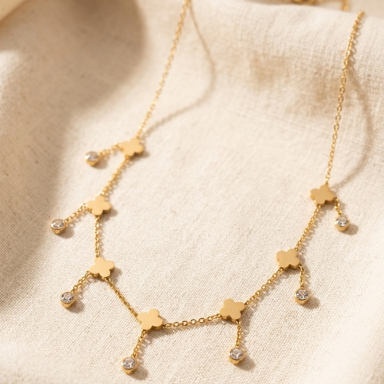A delicate gold necklace with small flower-shaped charms and clear rhinestone drops, displayed on a textured beige fabric background.