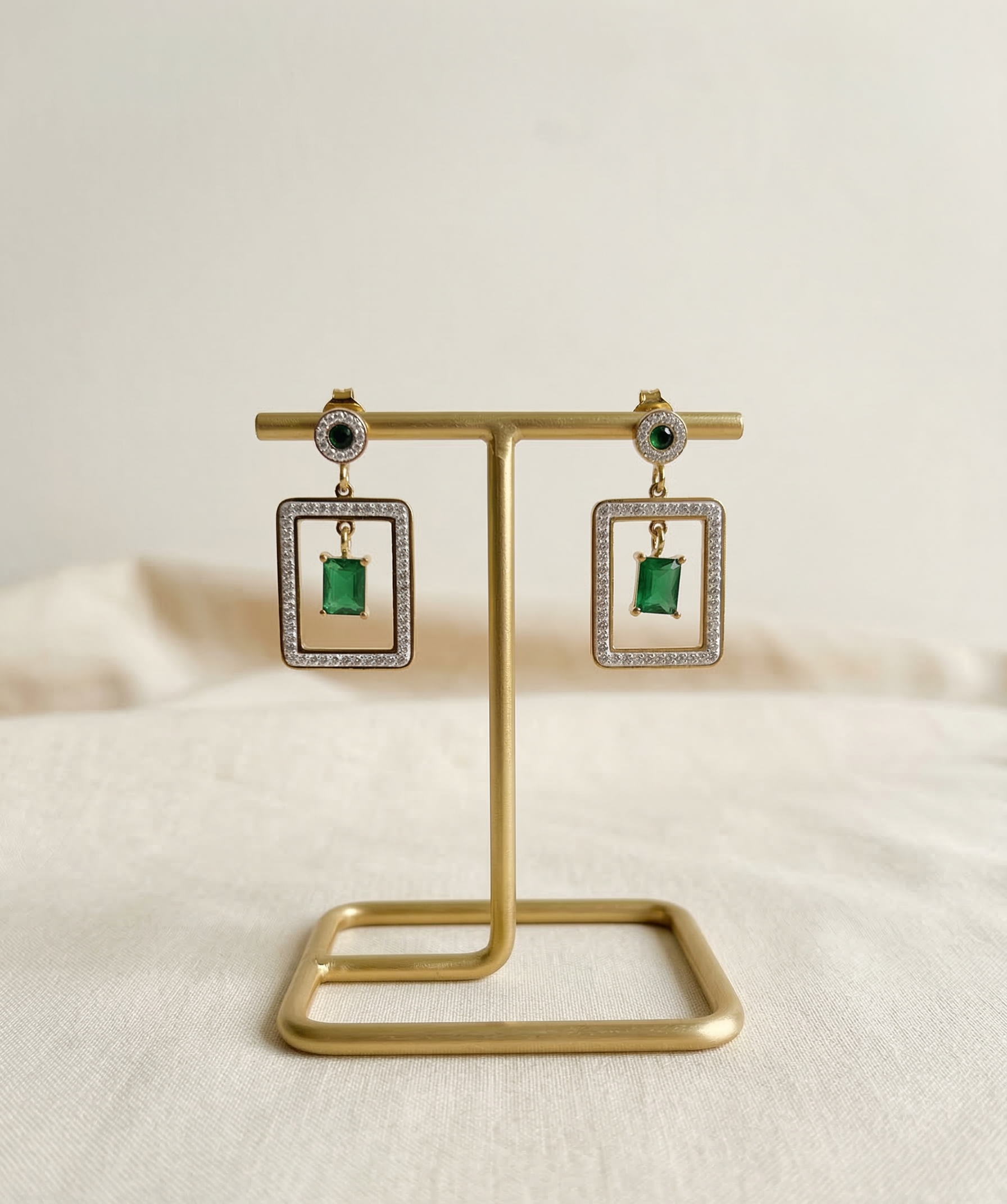 Pair of square earrings with green gemstones on a gold stand against a beige background