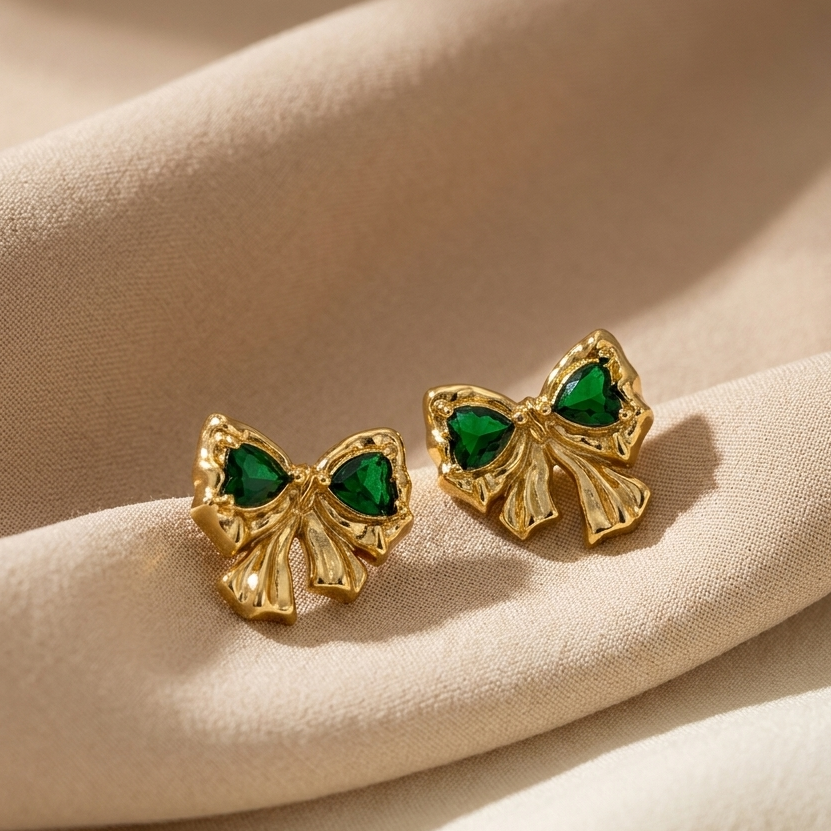 Gold bow-shaped earrings with green gemstones on a beige fabric background