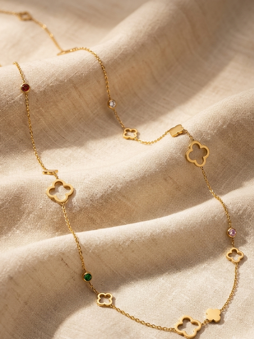 A delicate gold chain necklace featuring small openwork flower charms and tiny colorful gemstone accents, displayed on draped beige linen fabric.