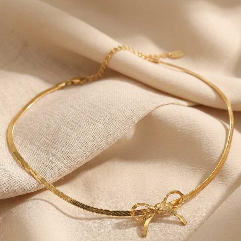 A delicate gold chain bracelet with a small bow charm, displayed on draped beige fabric.