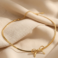 A delicate gold chain bracelet with a small bow charm, displayed on draped beige fabric.