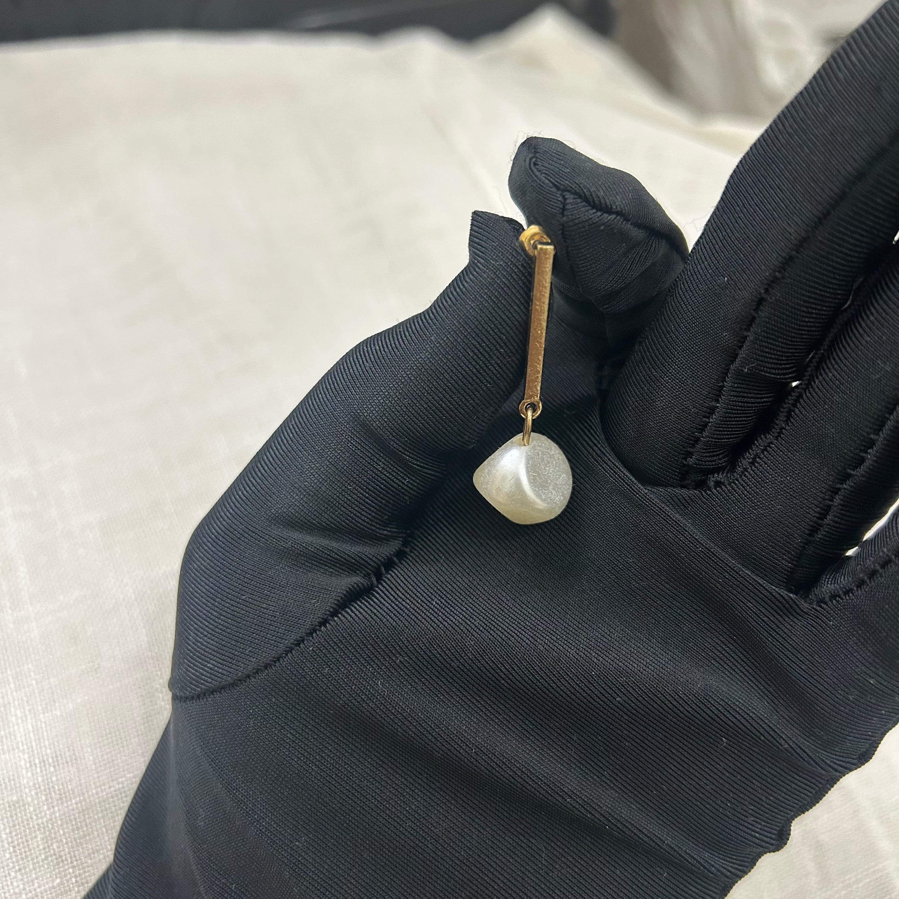 Pearl earring held by a gloved hand against a neutral background