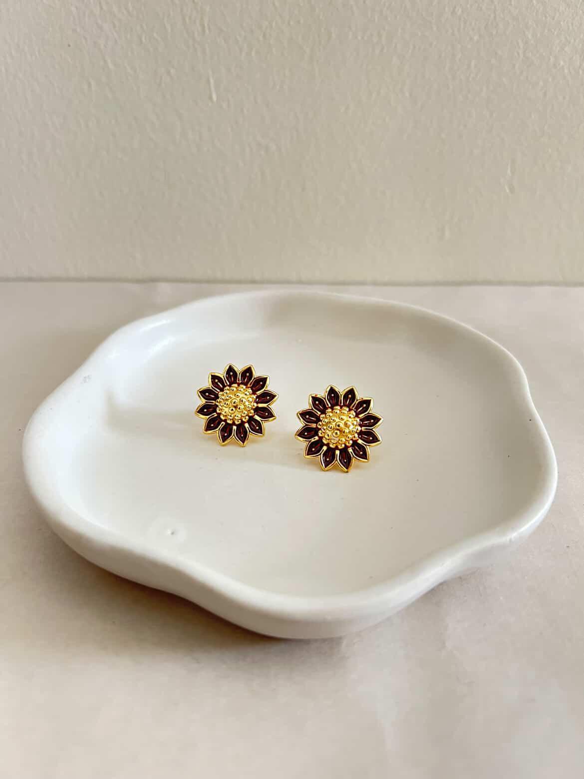 Gold and purple flower-shaped earrings on a white ceramic dish against a beige background