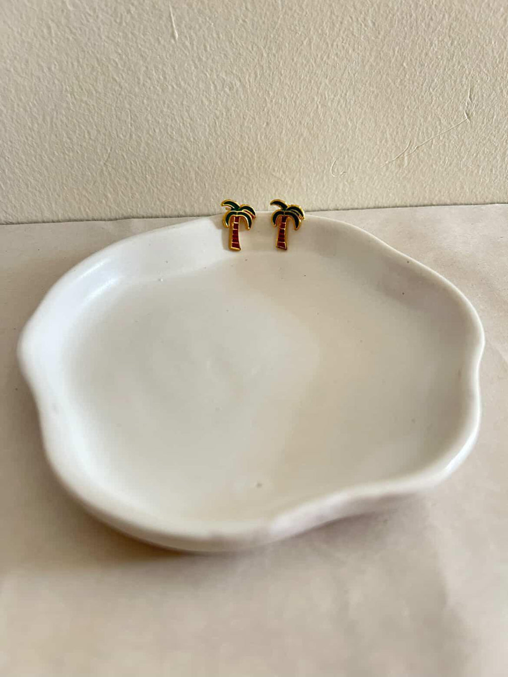 White ceramic dish with gold palm tree earrings on a beige surface.