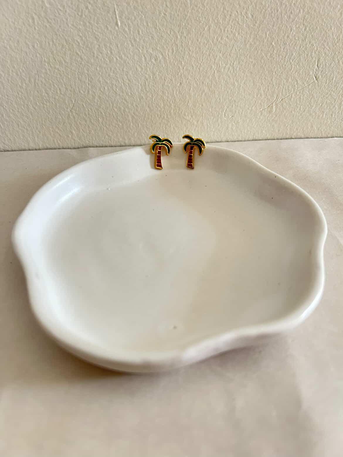 White ceramic dish with gold palm tree earrings on a beige surface.