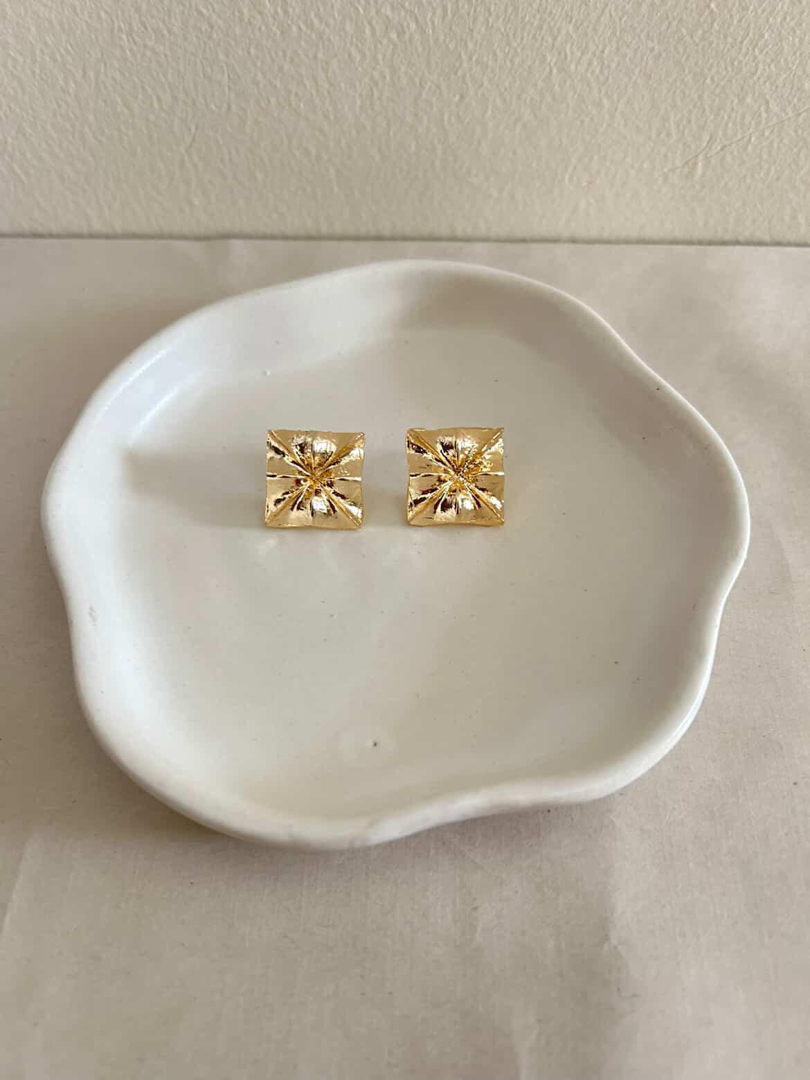 Gold square stud earrings on a white ceramic dish.