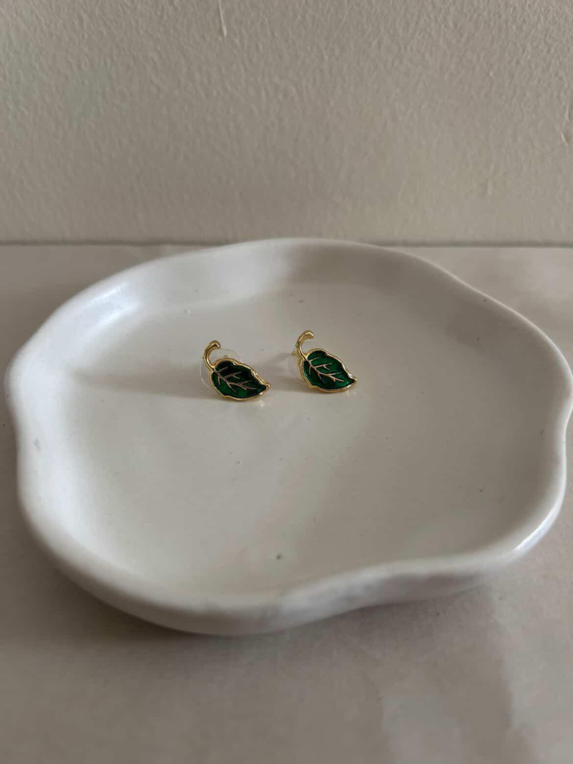 Gold and green leaf-shaped earrings on a white dish against a plain background