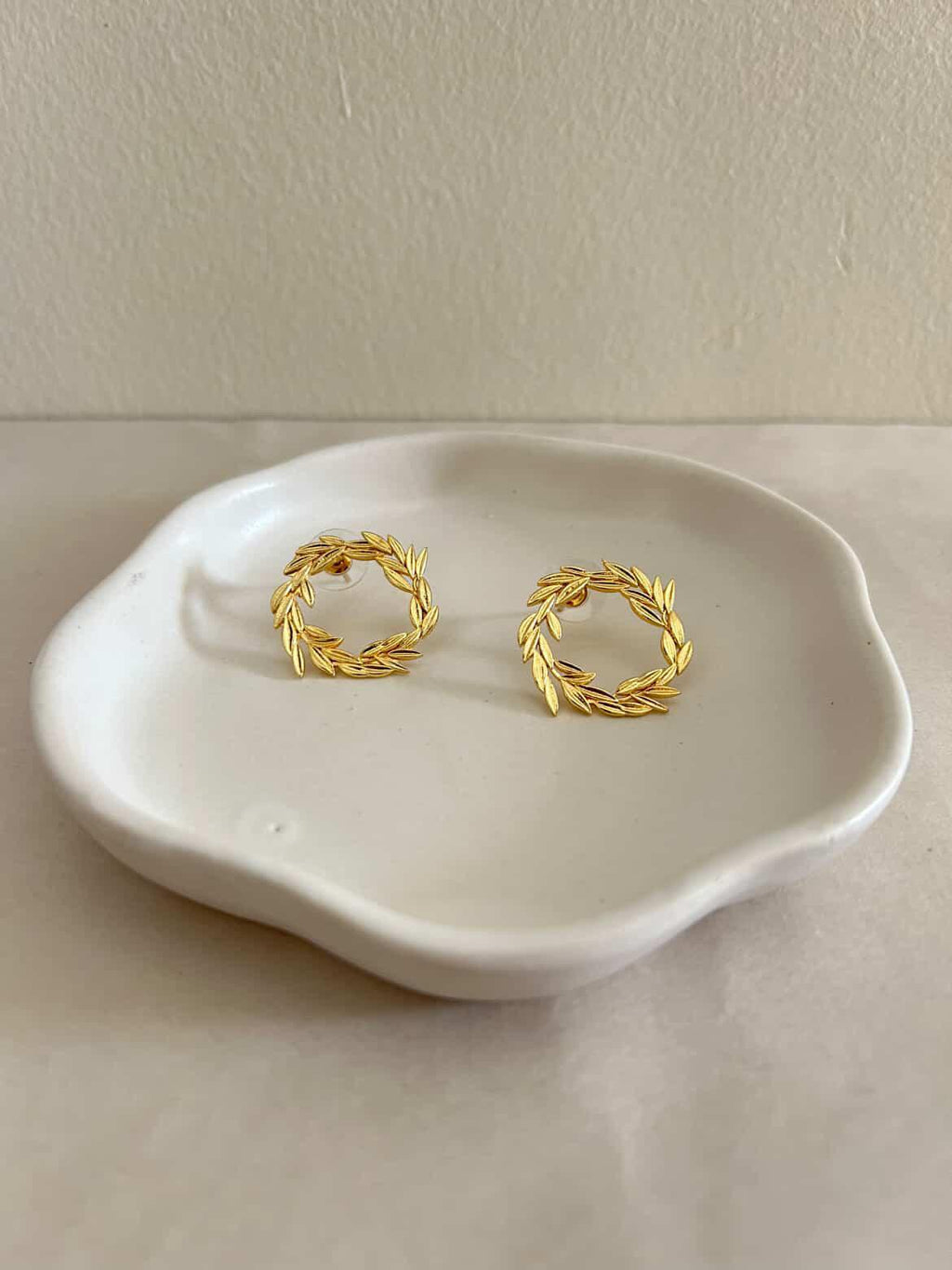 Gold wreath earrings on a white ceramic dish against a beige background