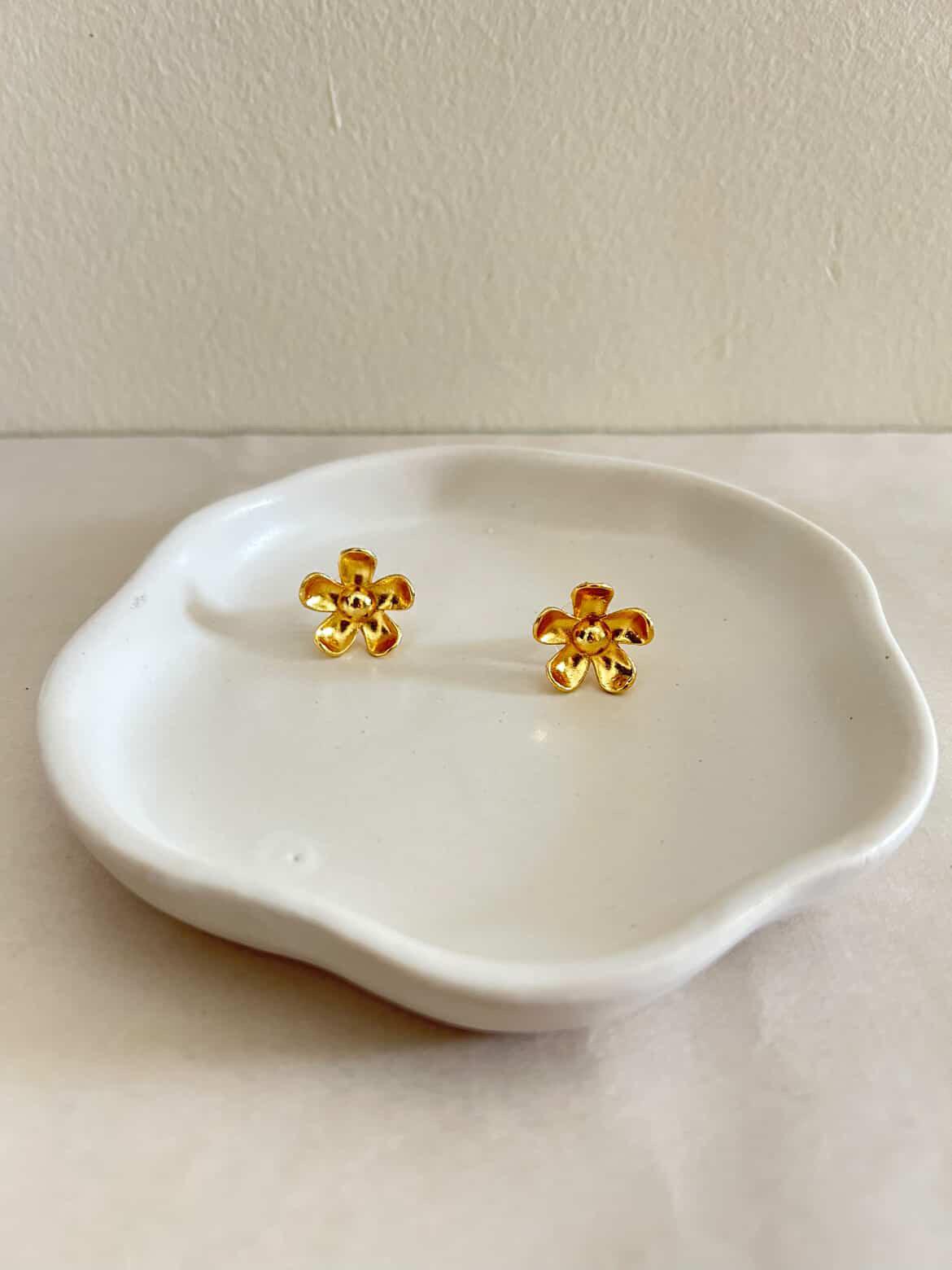 Gold flower-shaped earrings on a white ceramic dish against a beige background