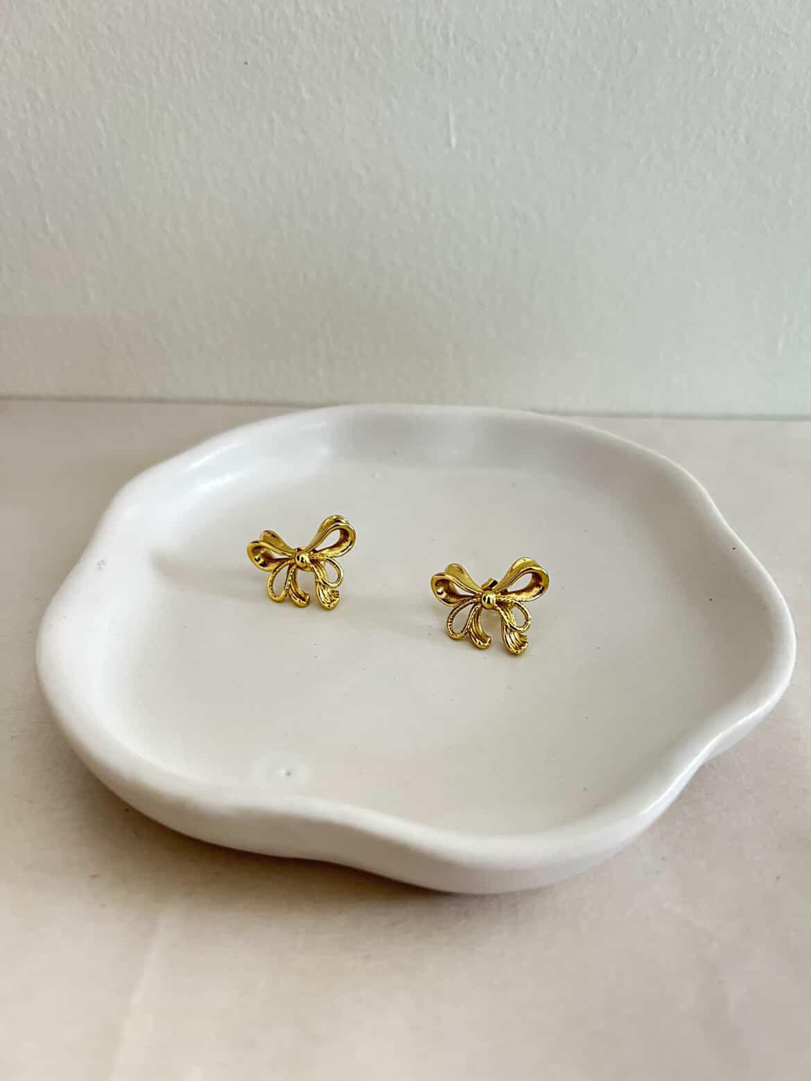 Gold bow-shaped earrings on a white ceramic dish against a light background