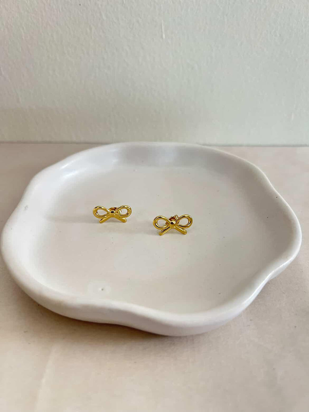 Gold bow-shaped earrings on a white ceramic dish against a plain background