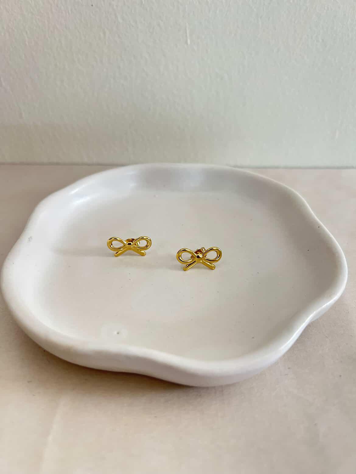 Gold bow-shaped earrings on a white ceramic dish against a plain background