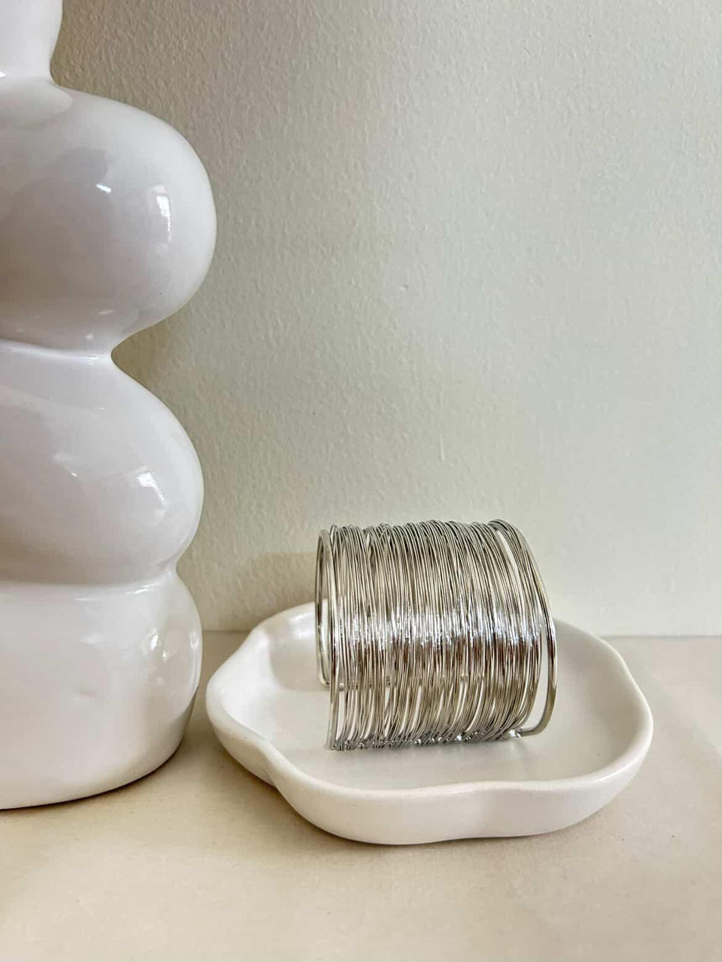 Stack of silver bracelets on a white dish next to a white vase against a light background